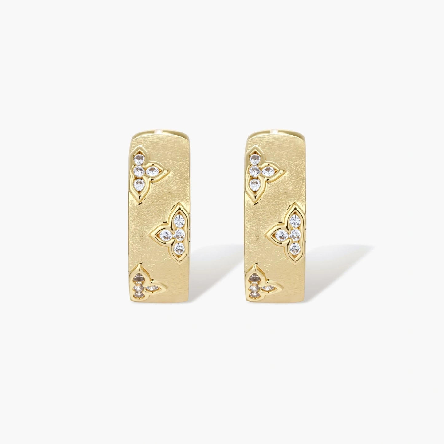 Gold-plated hoop earrings with textured surface and raised floral motifs set with clear zirconia stones on cream background.